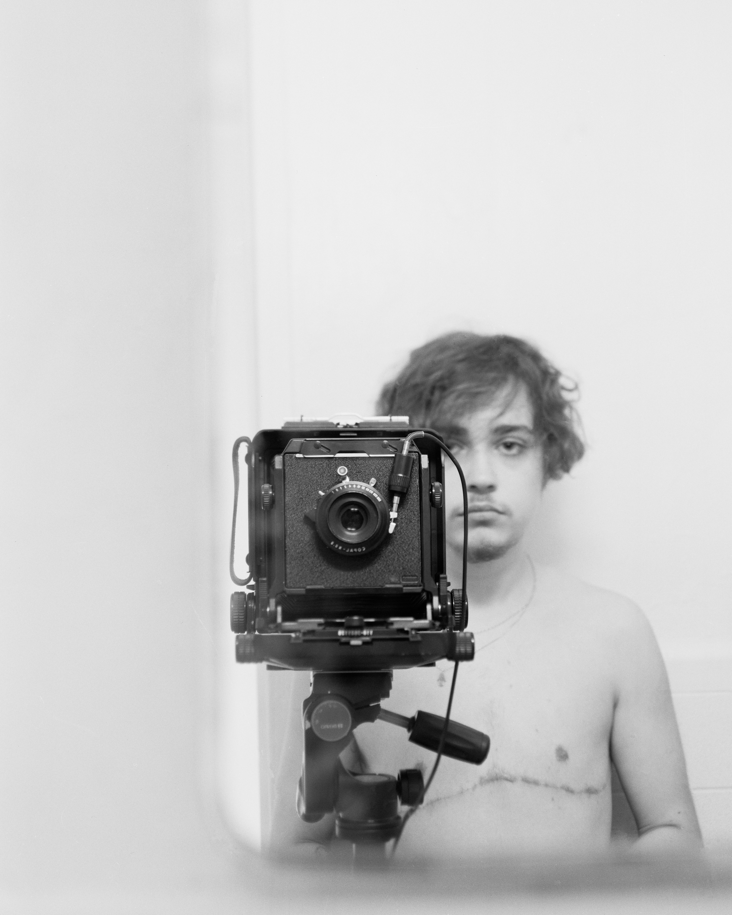 black and white mirror self portrait of the artist with top surgery scars using a 4x5 large format camera